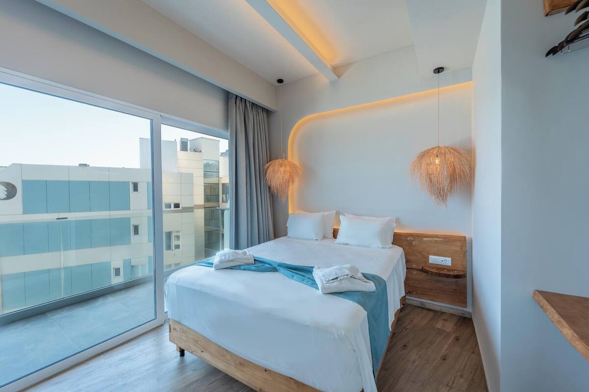 A modern hotel room with a large balcony, a bed with white linens, a wooden headboard, and two wall-mounted lamps.