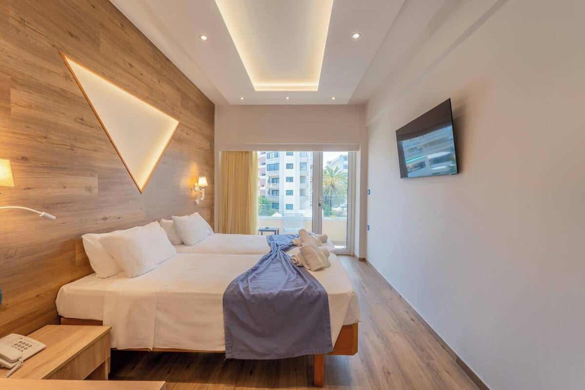 A hotel room with two beds, a wooden accent wall, a TV, and a balcony with a view of buildings and palm trees.