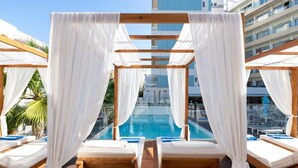 Zona de piscina con tumbonas de madera, cortinas blancas y vista despejada a la piscina.