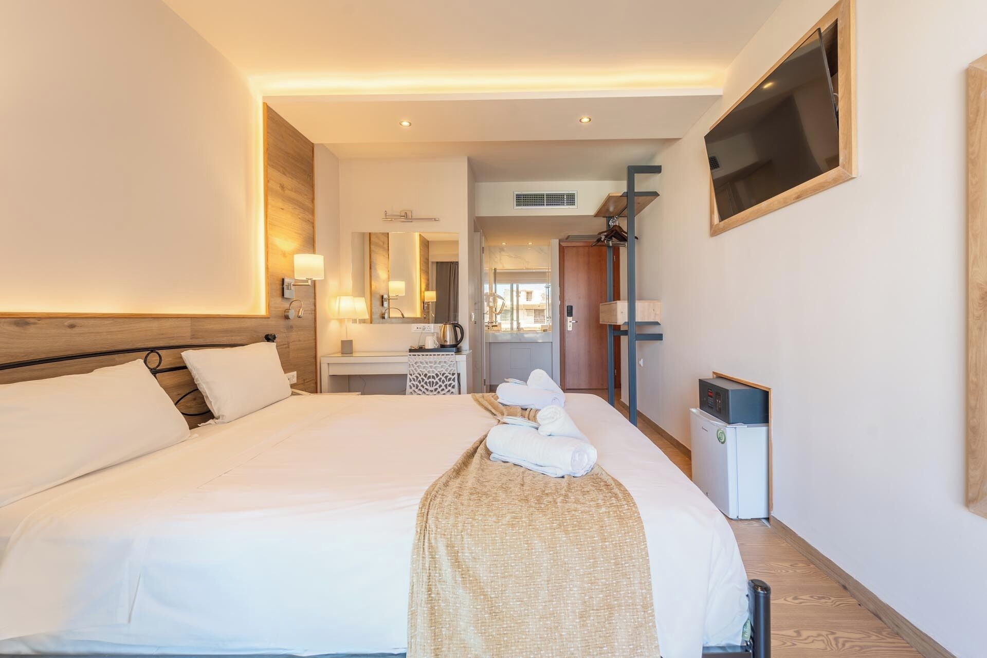 A hotel room with a large bed, a bedside table with a lamp, a mirror, a wardrobe, and a TV mounted on the wall.
