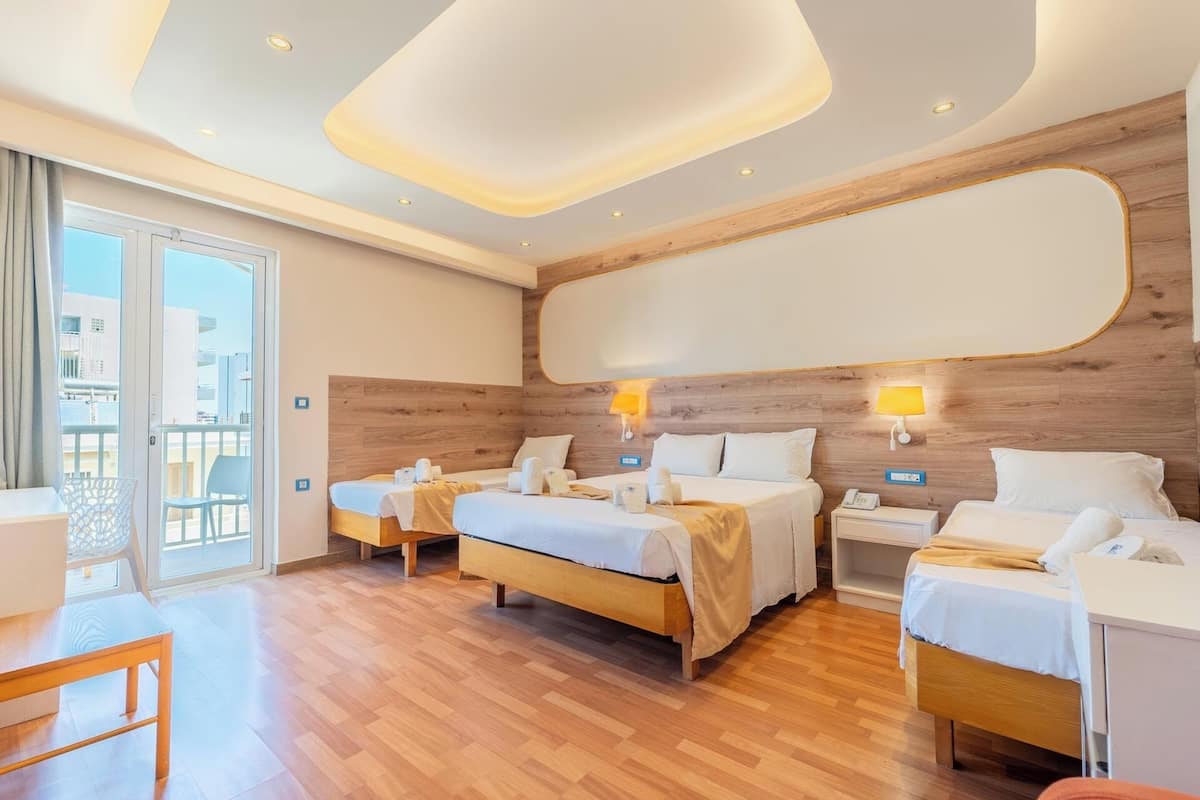 A hotel room with three beds, wooden flooring, a balcony with a view, and a modern ceiling design.