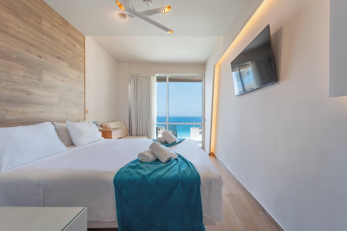 A hotel room with a large bed, a TV mounted on the wall, and a balcony with a view of the sea.