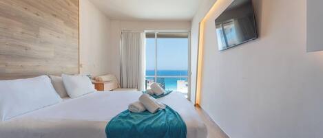 Habitación de hotel con una cama grande, un televisor montado en la pared y un balcón con vista al mar.