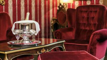 A room with a table set with a silver tray, two glasses, and a bottle. There are red velvet chairs and a wooden table with gold accents.