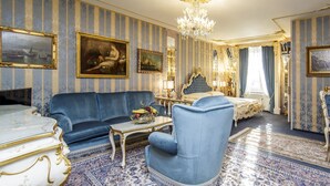 A room with a blue sofa, a white table, and a blue armchair. There are paintings on the walls and a chandelier. - Hotel Palais Porcia (Klagenfurt am Woerthersee)