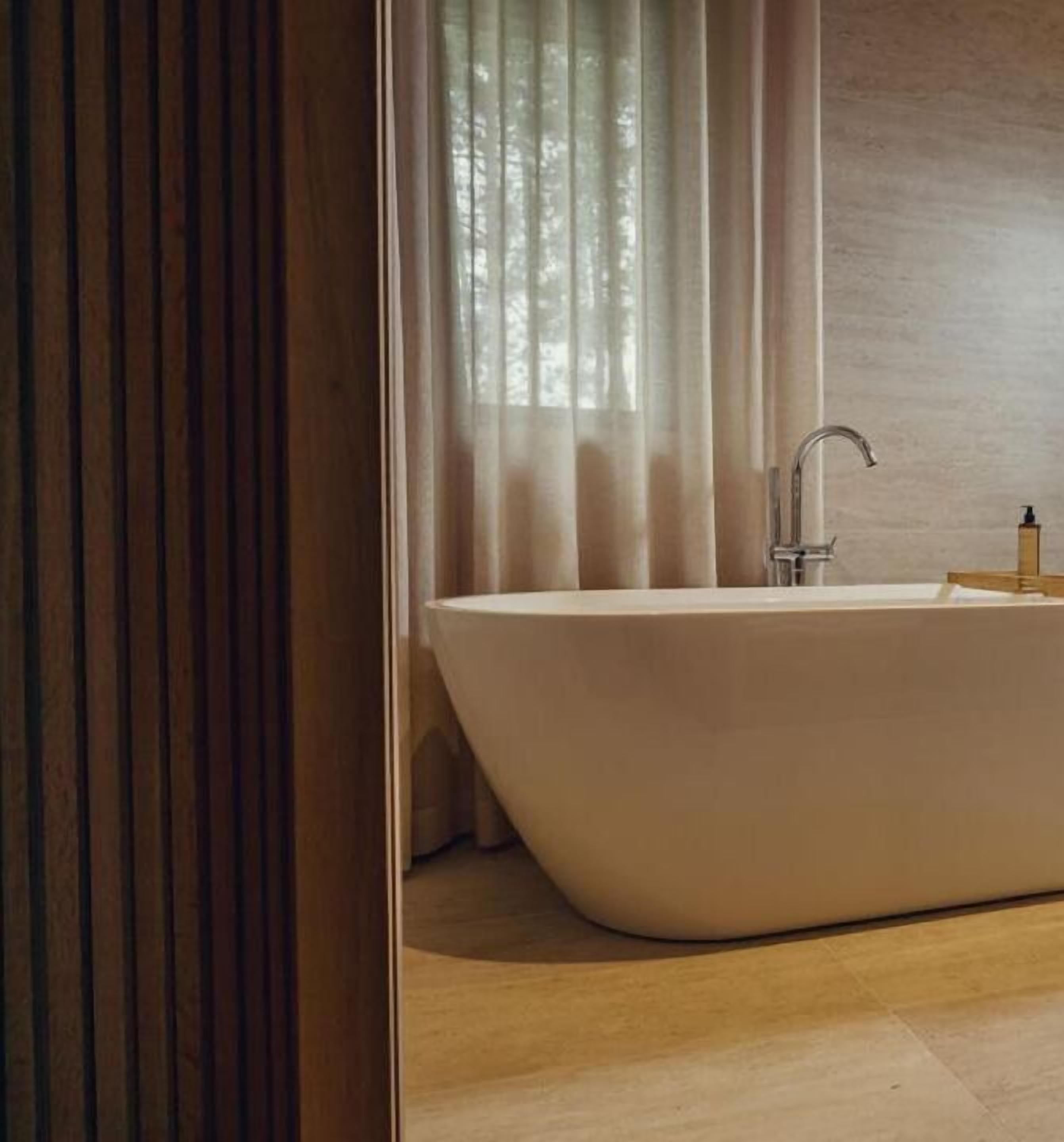 A modern bathroom with a freestanding bathtub, a wooden floor, and a window with sheer curtains.
