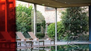 Un área de piscina interior moderna con sillones de descanso y un muro de cristal que refleja la vegetación circundante.