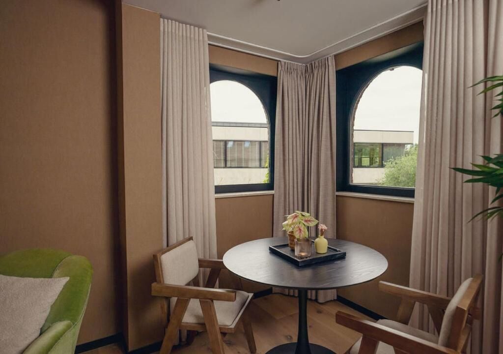 Een kamer met een ronde tafel, twee stoelen, een potplant en grote ramen met gordijnen.