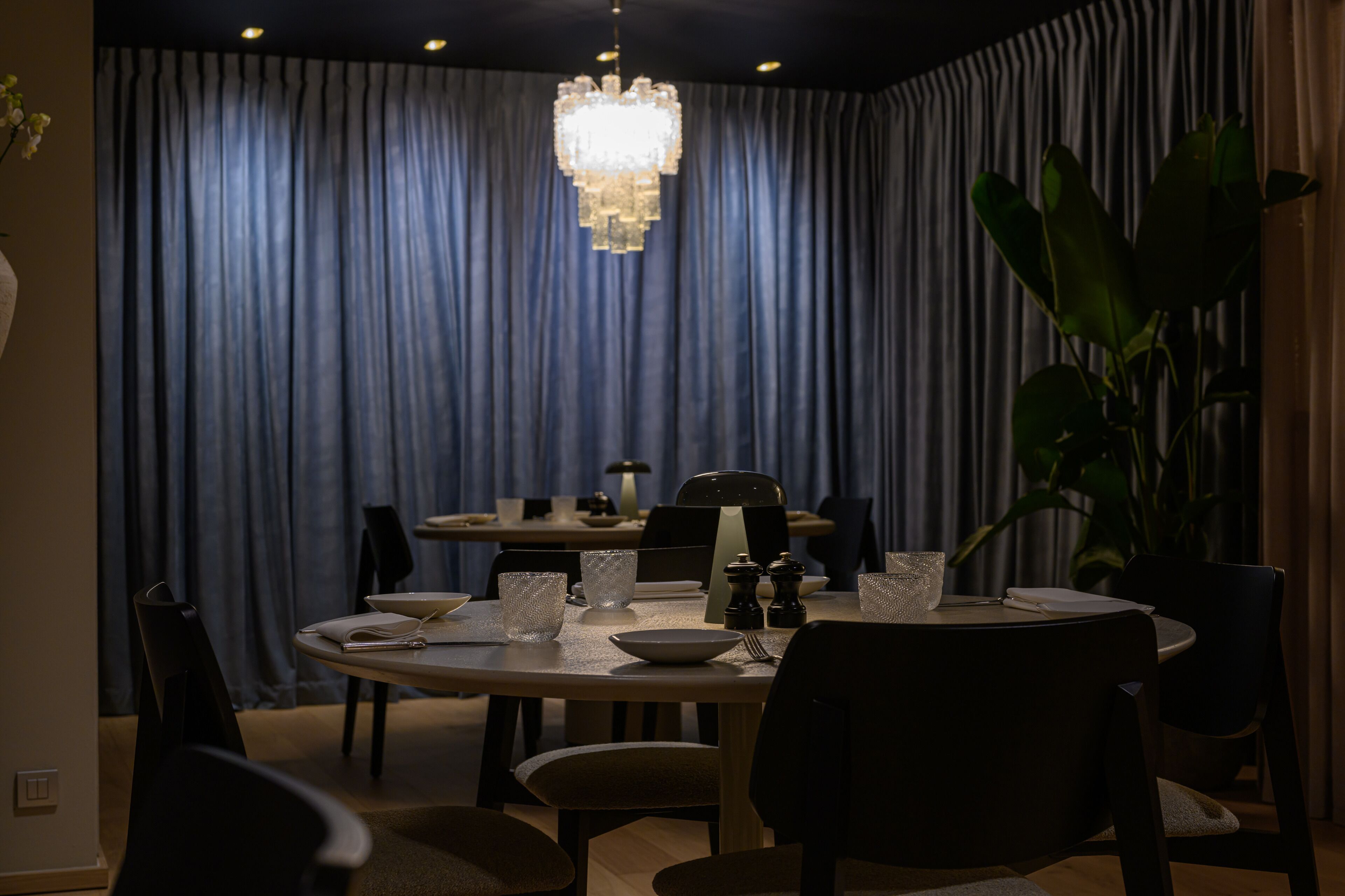Een eetgedeelte met een ronde tafel, stoelen, een kroonluchter en een gordijnen achtergrond.