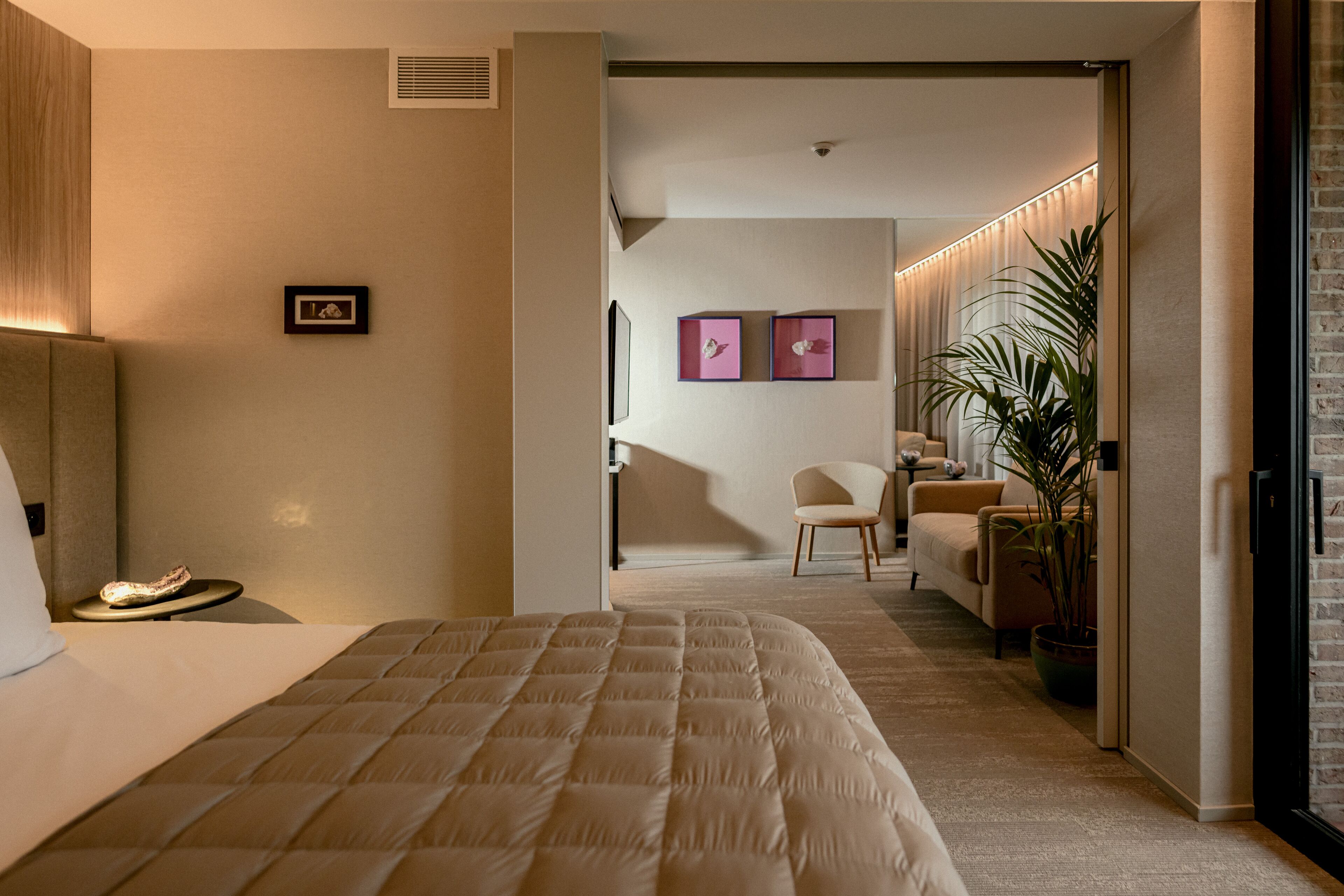Una habitación de hotel moderna con una cama grande, una zona de estar con sofá y sillones, un televisor de pantalla plana y una planta en maceta.