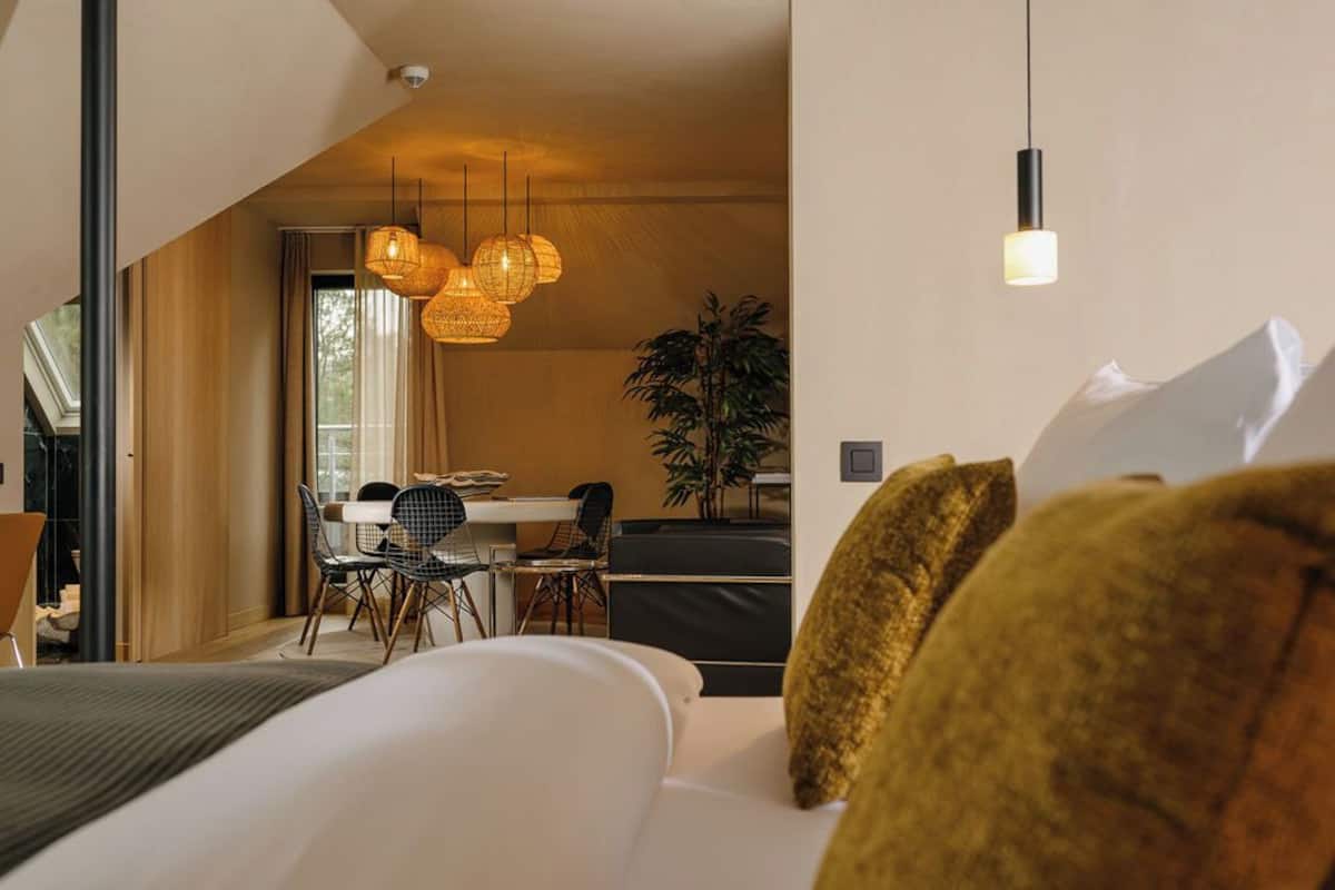 A modern hotel room with a large bed, a dining area, and a view of the outdoors.