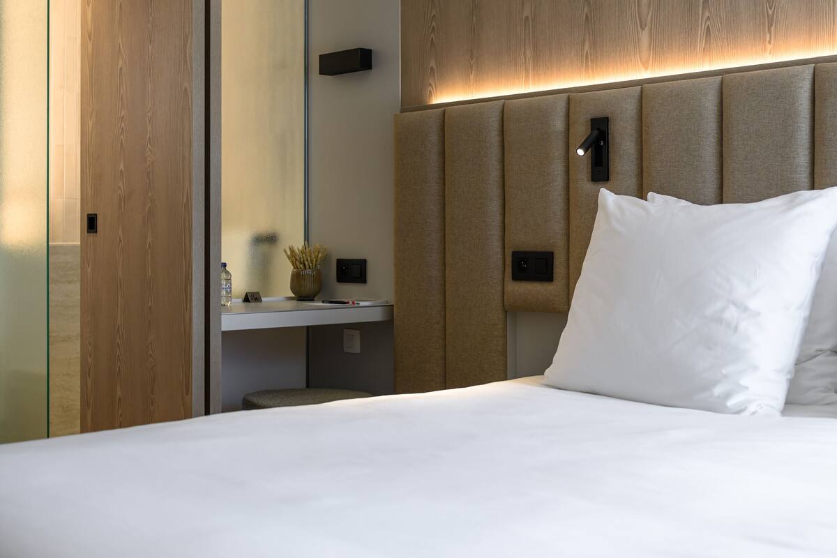 A modern hotel room with a wooden headboard, a large bed with white linens, a bedside table with a vase, and a mirror on the wall.