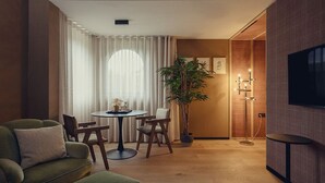 Una sala de estar con un sofá verde, una mesa redonda con sillas, una planta en maceta y un televisor en la pared.