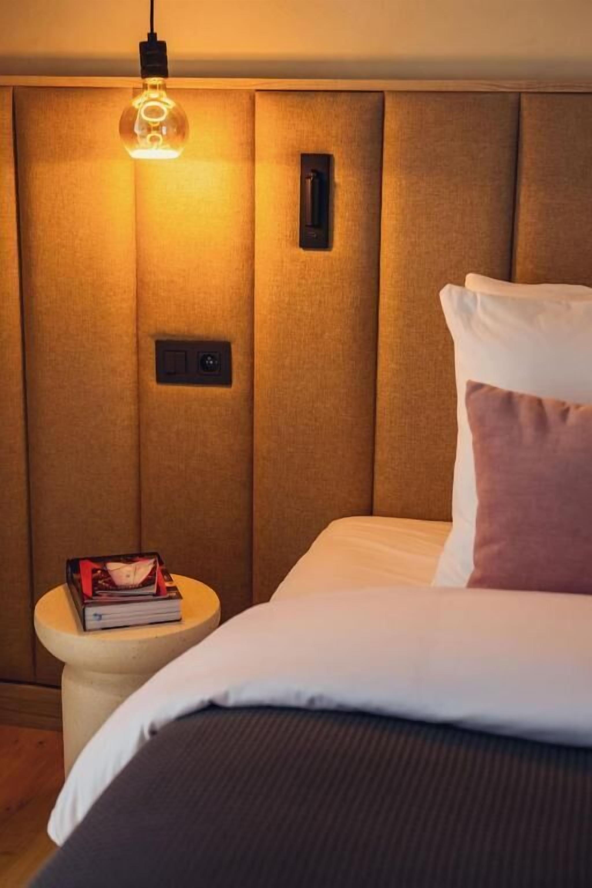 Une chambre d’hôtel avec un lit, une table de chevet sur laquelle se trouve un livre, une lampe fixée au mur et un panneau de commande.