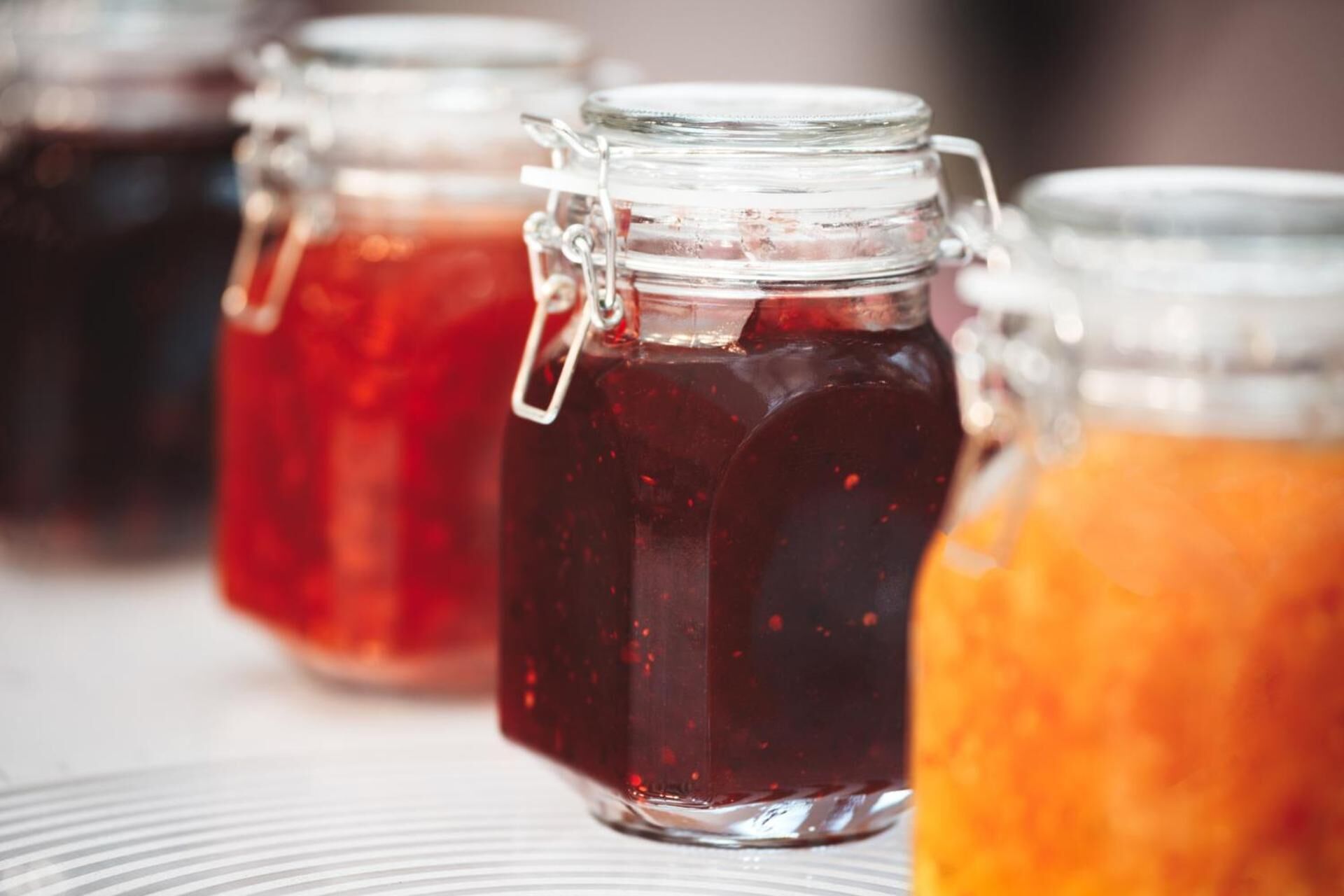 Een rij glazen potten met metalen sluitingen, gevuld met vloeistoffen in verschillende kleuren, waarschijnlijk zelfgemaakte jam of sauzen.