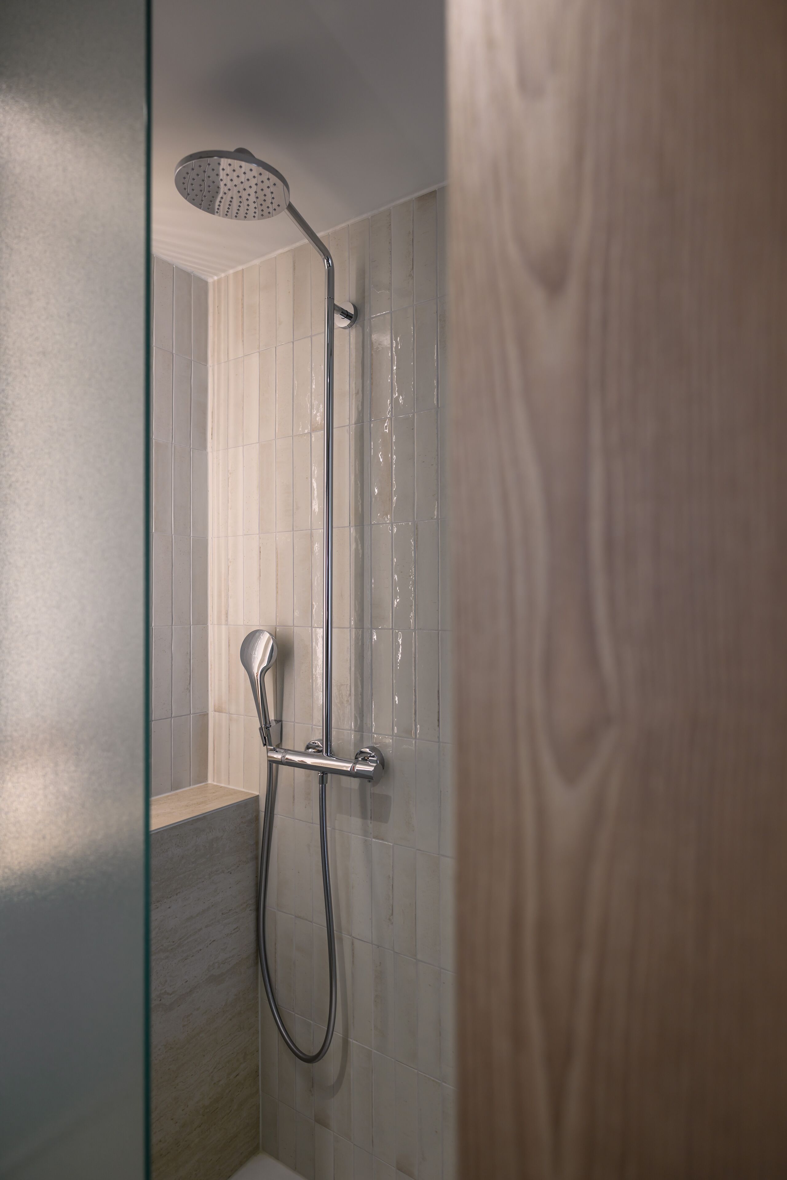 Ducha con cabezal de ducha y manguera de mano, frente a un pared de azulejos con un panel de madera.