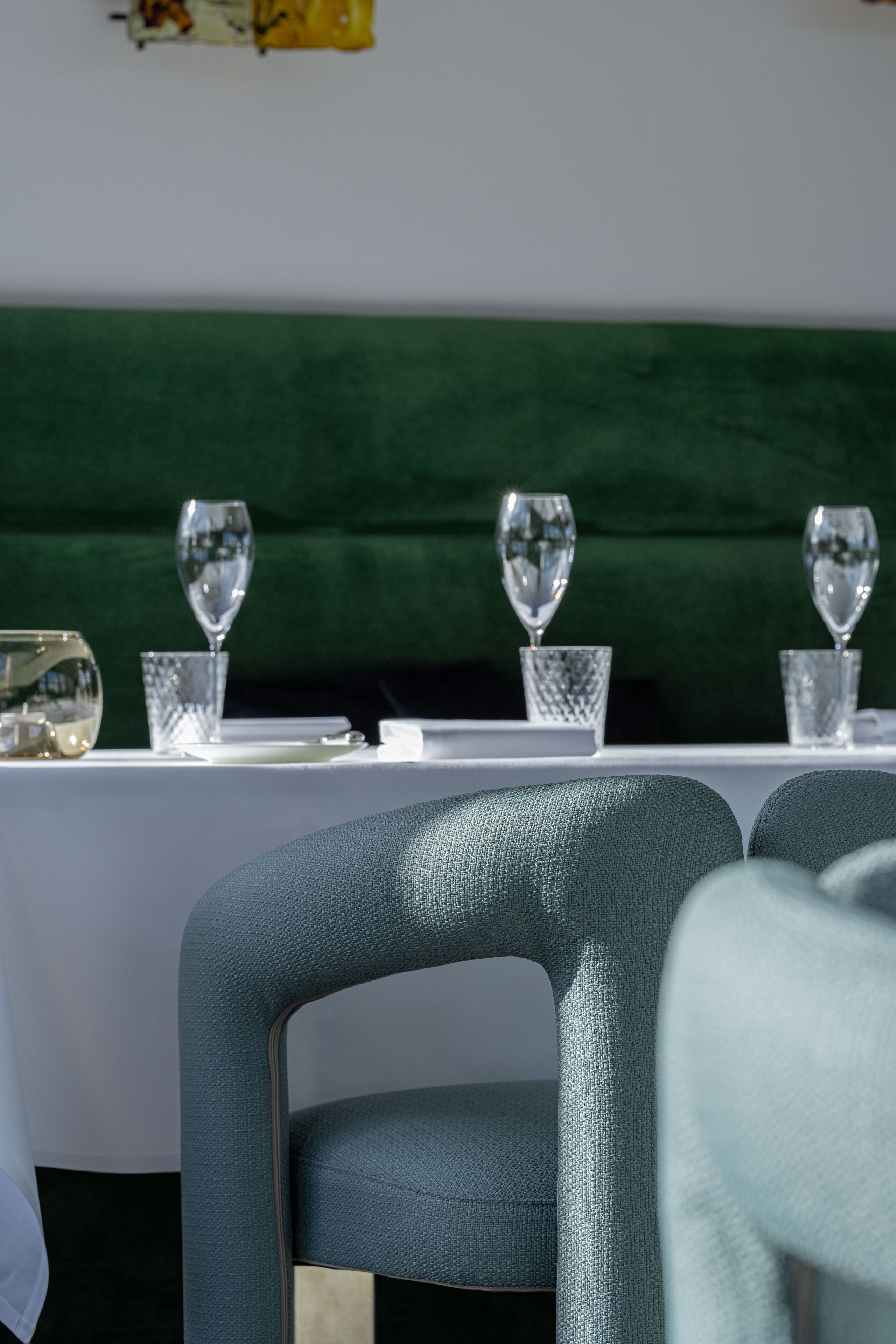 A dining table with a green upholstered headboard, clear glassware, and a white tablecloth.