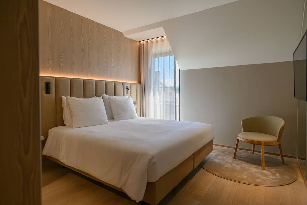 A modern bedroom with a large bed, a wooden headboard, a chair, and a view of the outdoors.