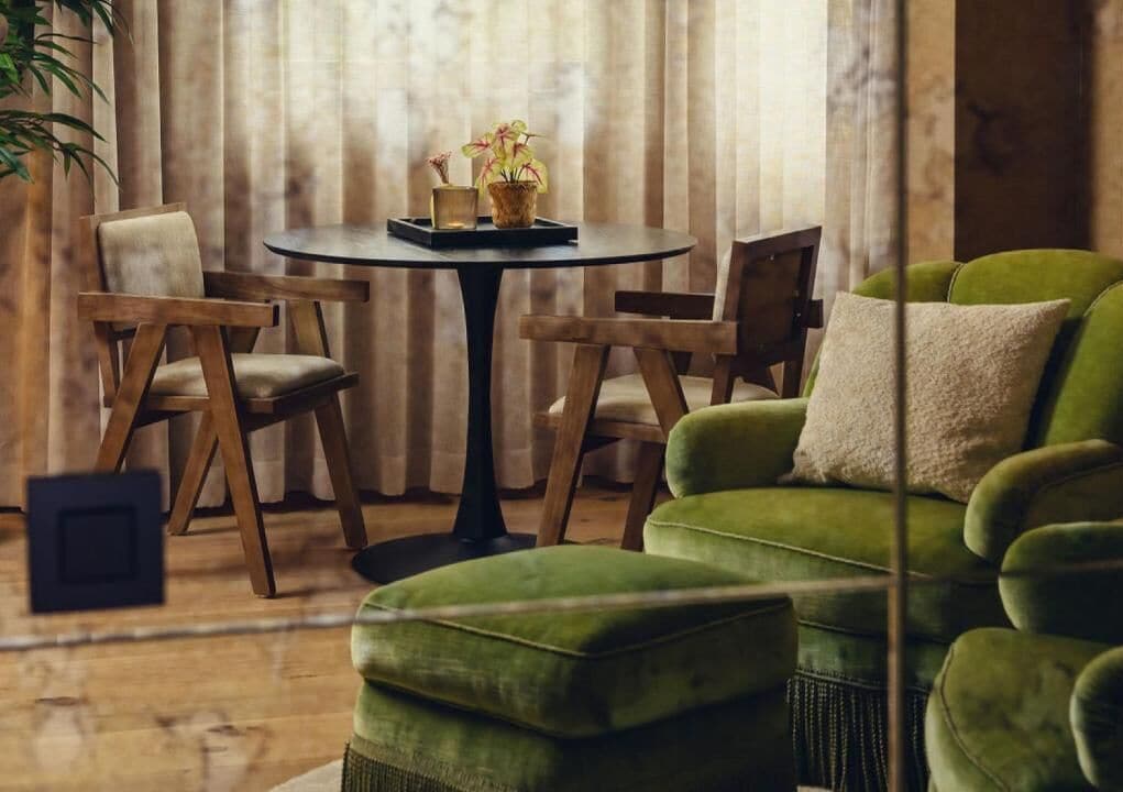 Una sala de estar acogedora con un sofá verde, dos sillas de madera, una mesita redonda con una bandeja y plantas en macetas.