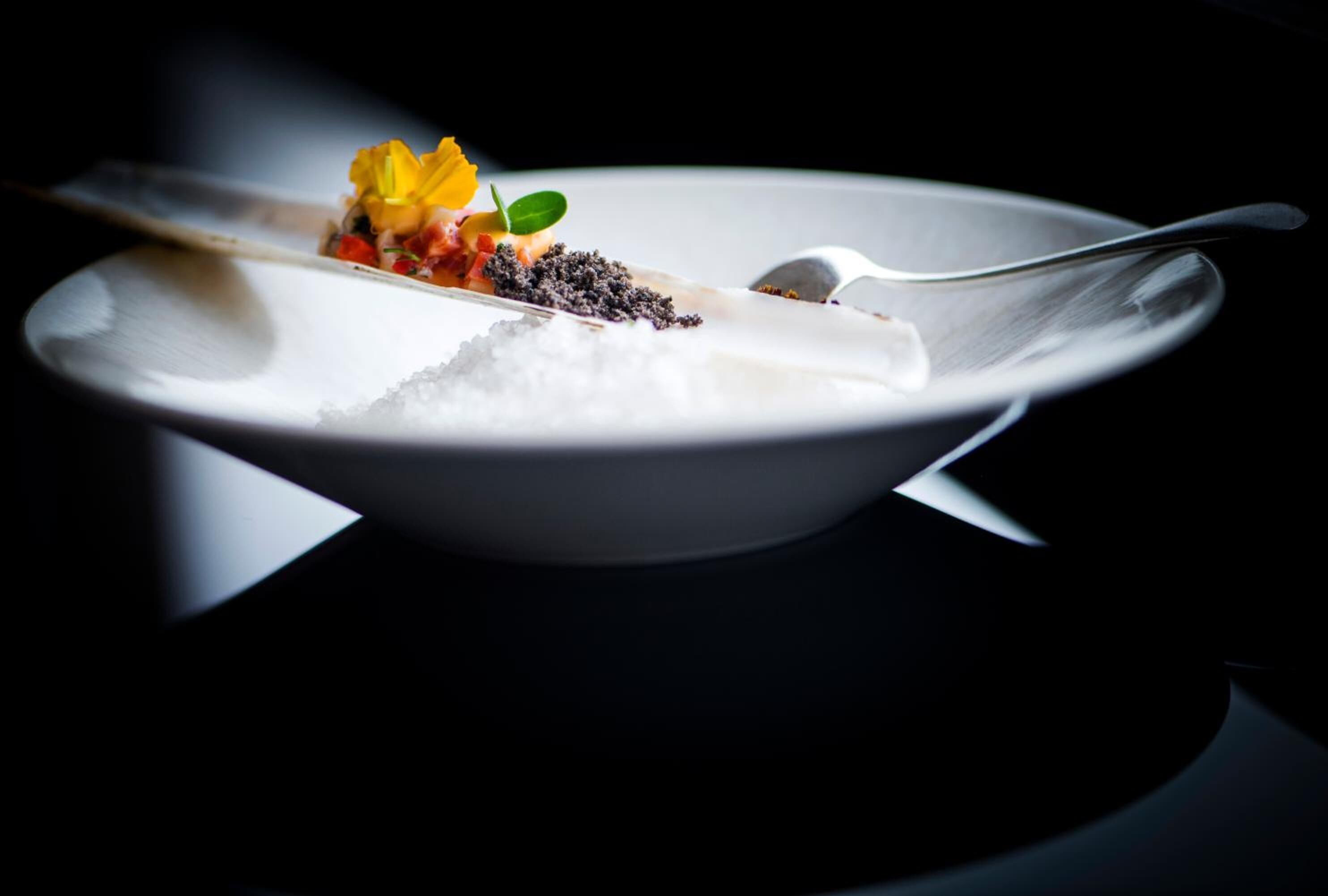 Un plato blanco con una cuchara, que contiene un plato con una flor amarilla, semillas negras y una sustancia blanca.