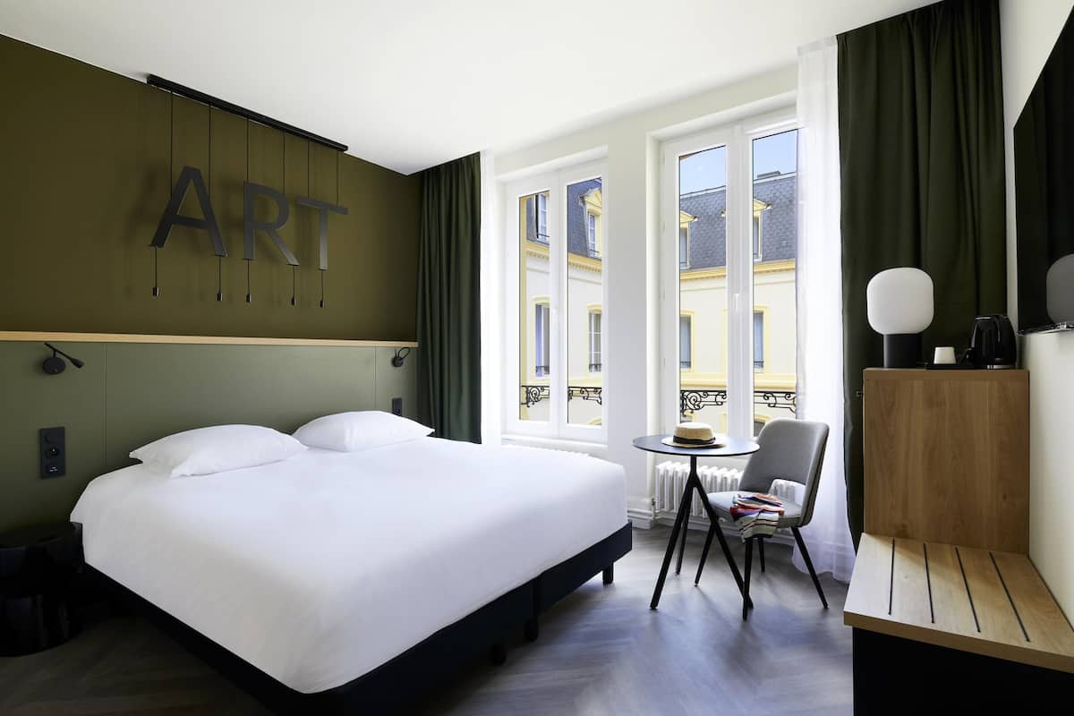 A hotel room with a bed, a small table, a chair, a desk, and a view of buildings through large windows.