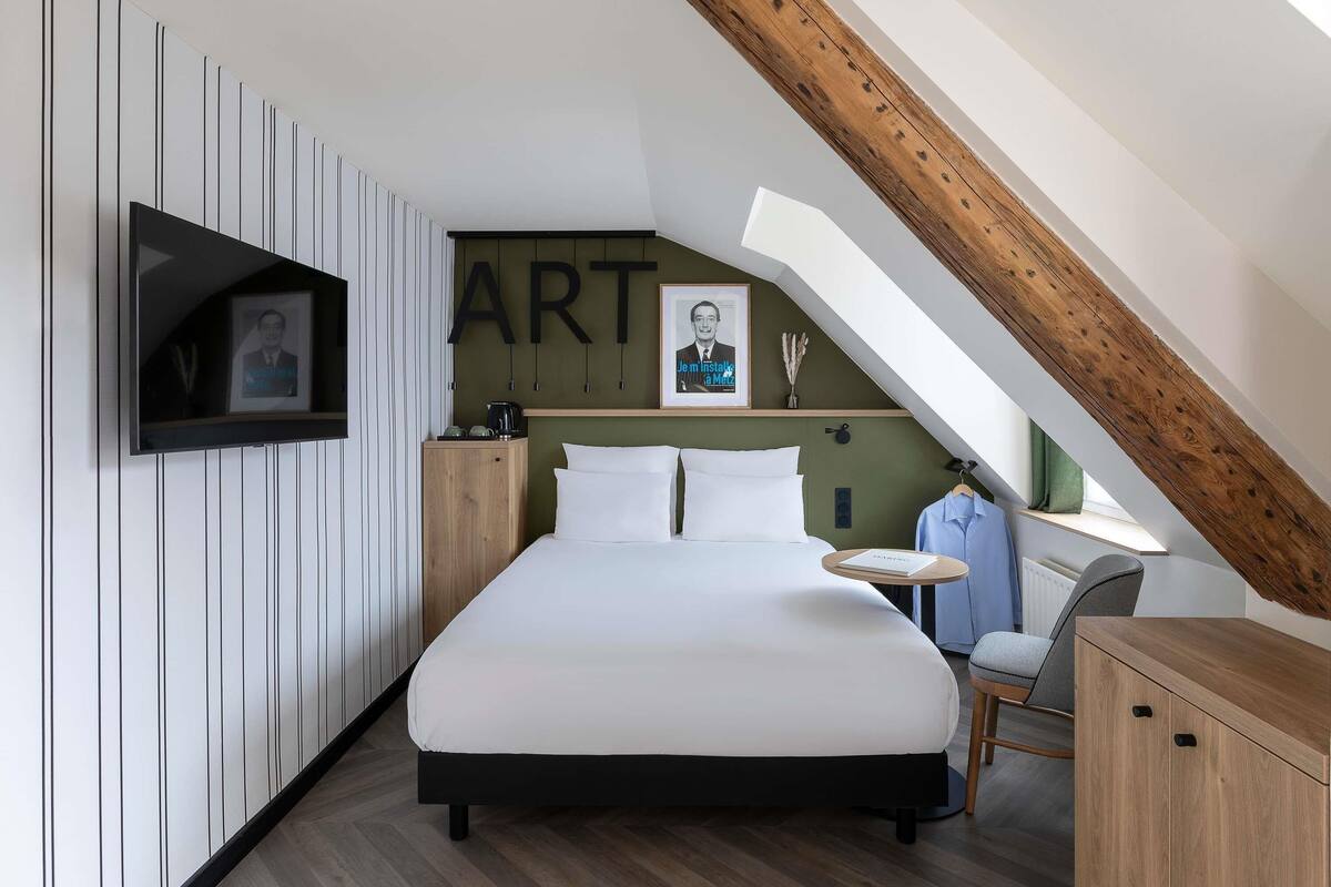 A modern bedroom with a sloped ceiling, a bed, a TV mounted on the wall, and a small table with a chair.
