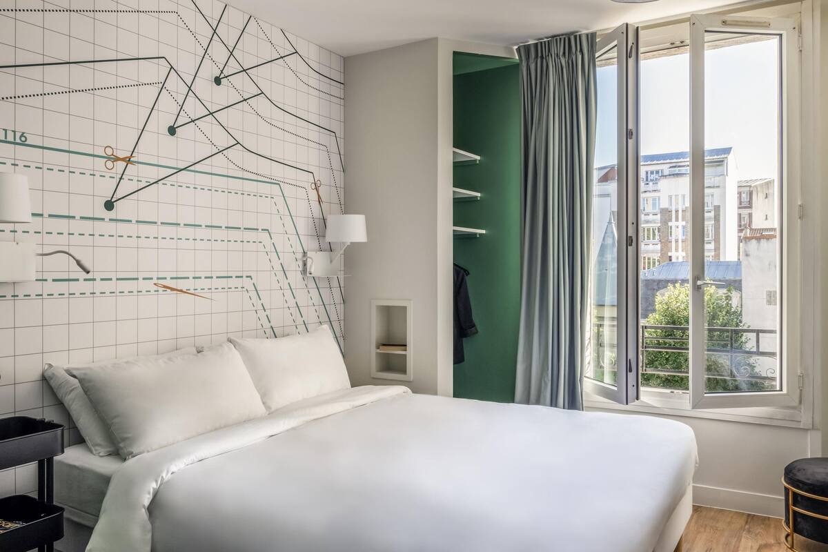 A modern bedroom with a large bed, a geometric wall design, a window with curtains, and a view of buildings outside.