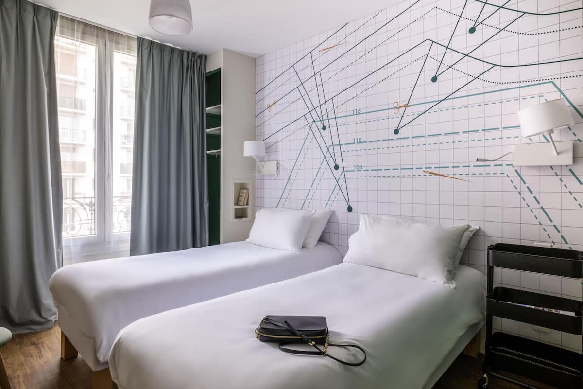A hotel room with two beds, a large window with curtains, a wall with a geometric design, and a black luggage cart.