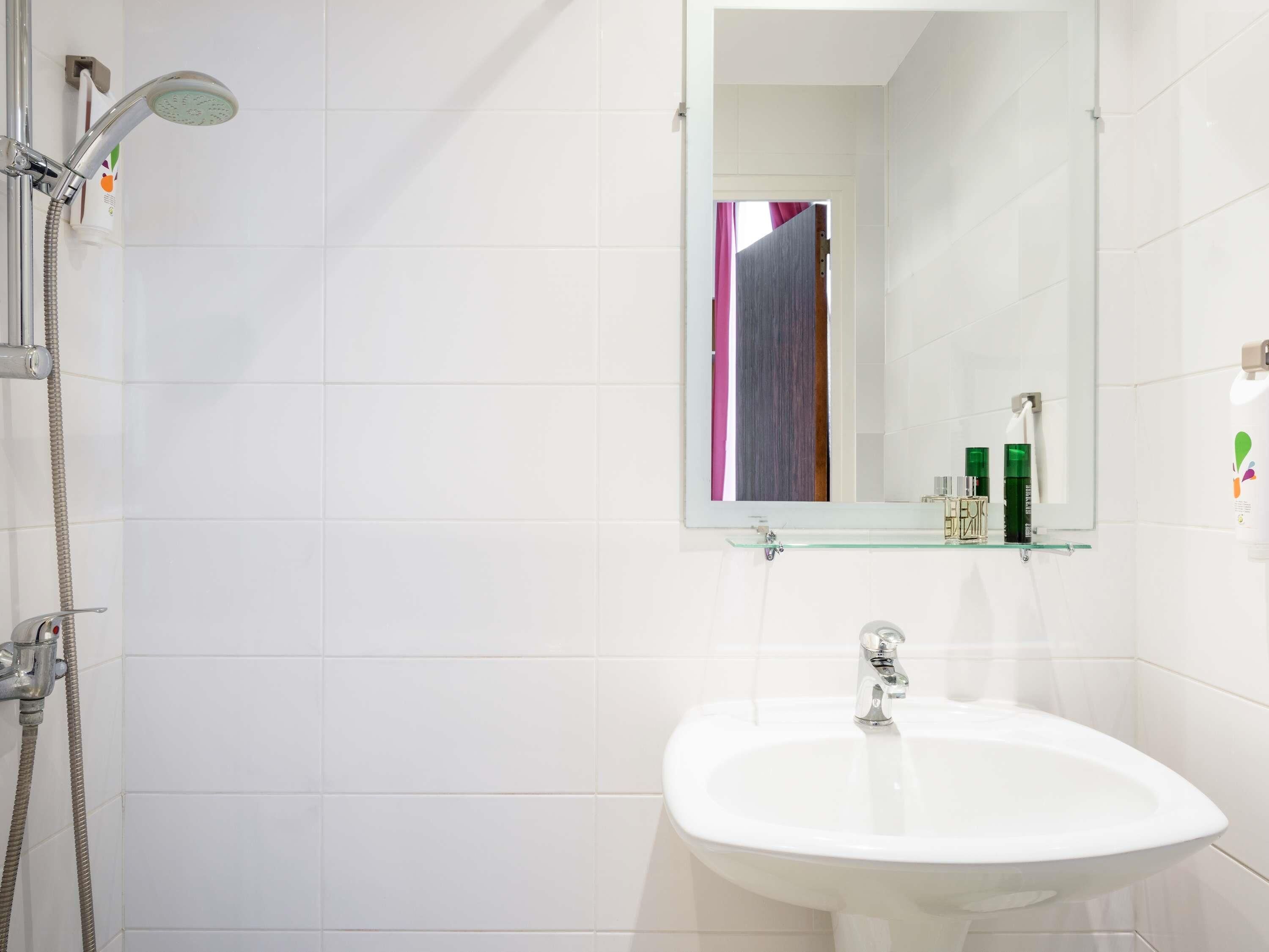 Une salle de bain avec un lavabo blanc, une étagère en verre où sont rangés les articles de toilette, une douche avec un pommeau de douche à main et un miroir qui donne sur une pièce séparée par un rideau.