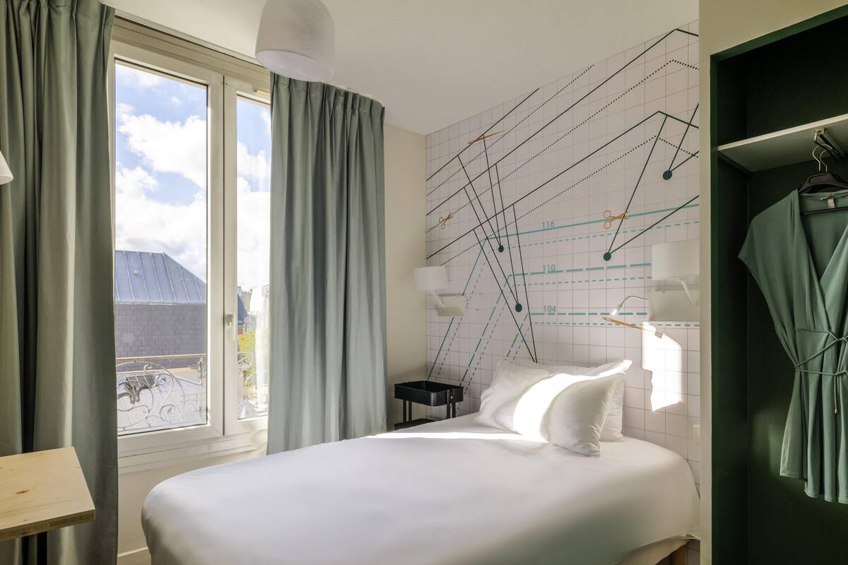 A bedroom with a bed, a nightstand, a lamp, a window with curtains, and a wall with a grid pattern.