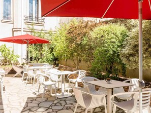 Patio esterno con sedie e tavoli in plastica bianca, un ombrello rosso e un pavimento in pietra.