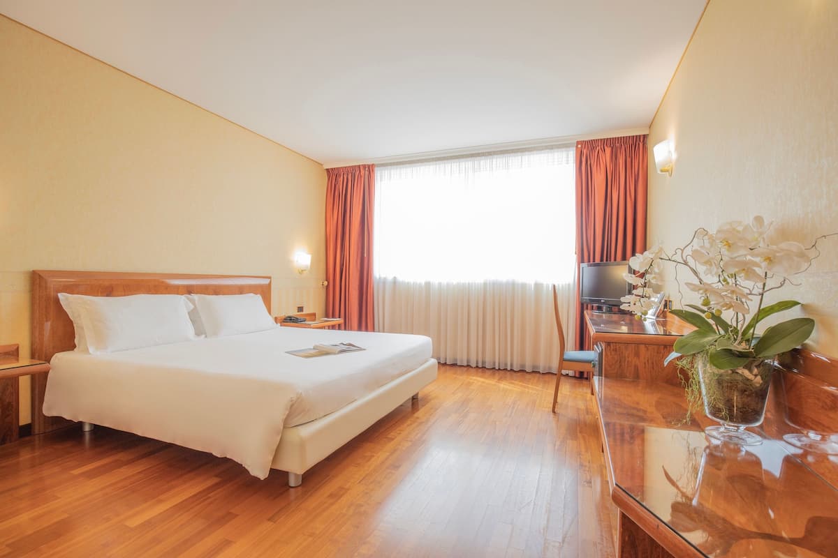 A hotel room with a wooden floor, a bed with white linens, a desk with a TV, and a potted orchid.