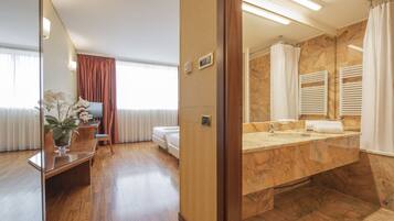 A modern hotel room with a wooden floor, a sitting area with a plant, a bed, and a bathroom with a marble sink and towel racks.