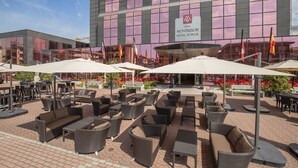 Un patio al aire libre con mobiliario de mimbre y sombrillas, frente a un edificio de hotel con fachada de vidrio moderno.