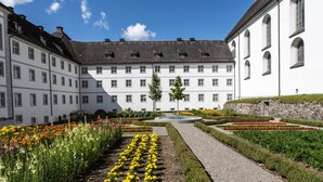 Un patio con una fuente central, rodeado de diversos macizos de flores y un edificio grande con múltiples ventanas y una pared de piedra.