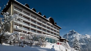 Un hotel de varios pisos con balcones, rodeado de árboles y montañas nevadas.