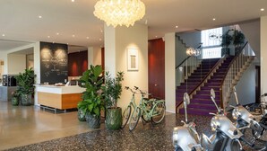 Um hall de hotel com balcão de recepção, plantas em vasos, bicicletas, scooters, escadaria e um lustre.
