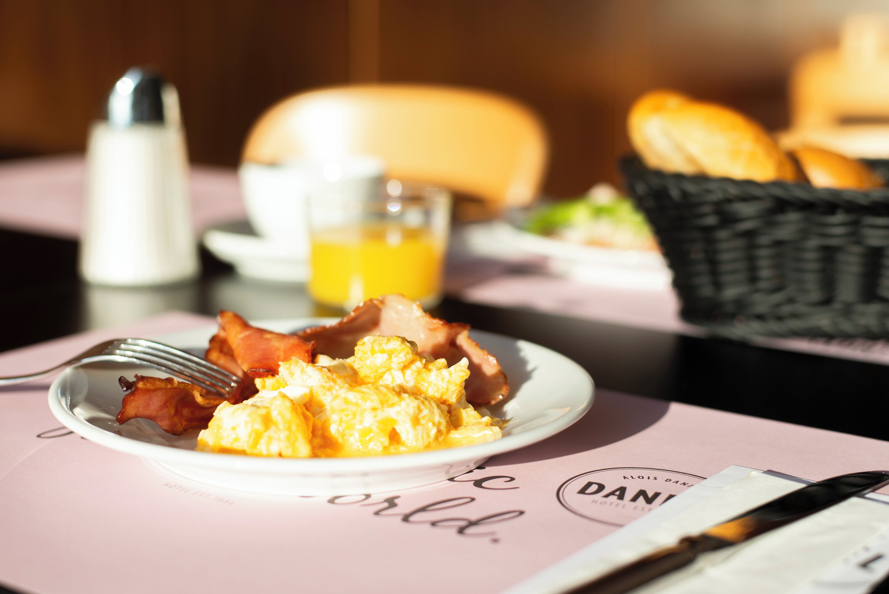 A breakfast setting with scrambled eggs, bacon, and toast on a plate, a glass of orange juice, and a basket of bread on a table.