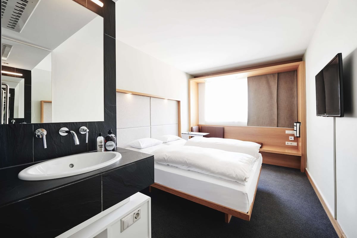 A modern hotel room with a large white sink, a mirror, two beds, a small bedside table, and a TV mounted on the wall.