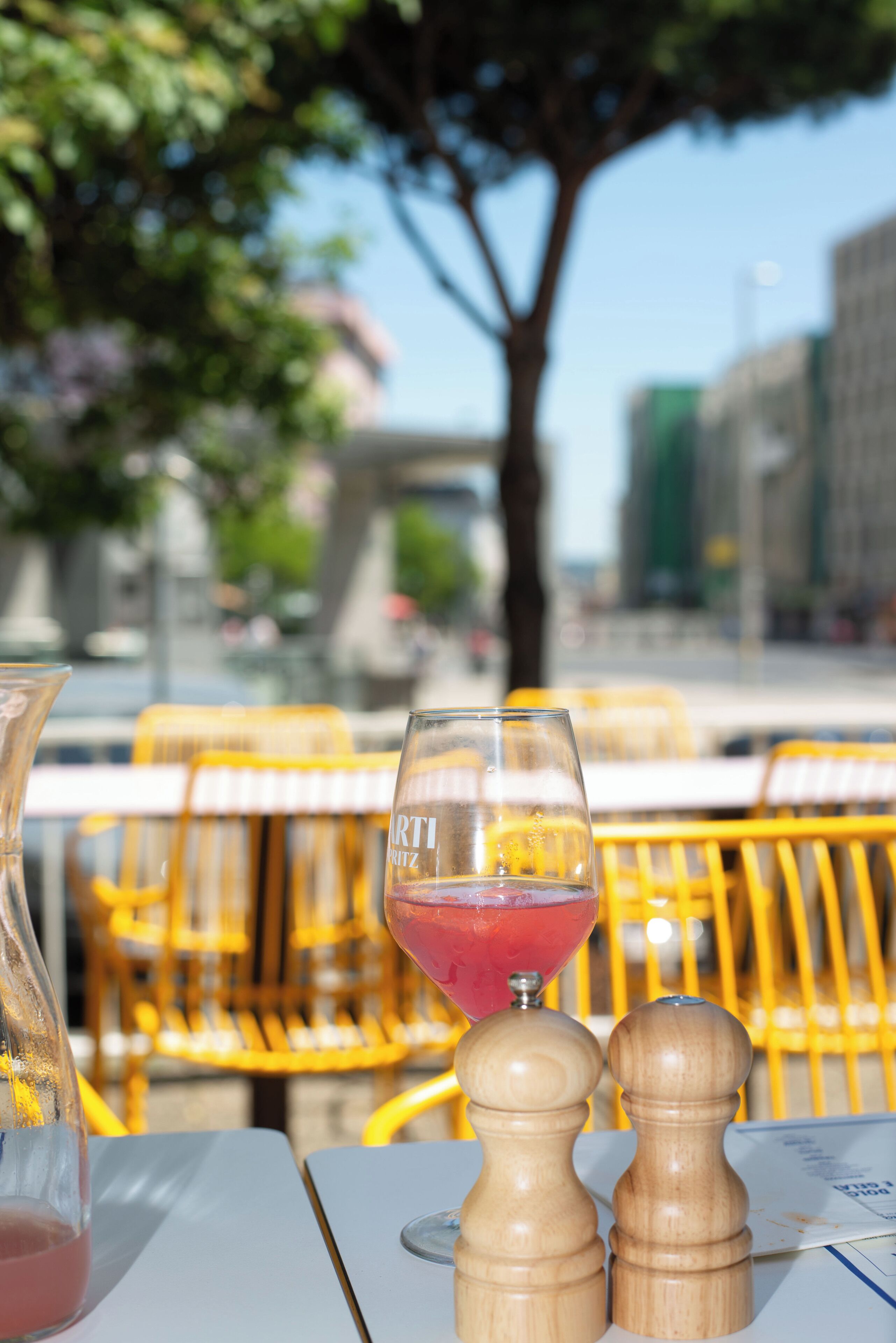 A table with a glass of red drink, salt and pepper shakers, and a pitcher, set outdoors with yellow chairs and a tree in the background.