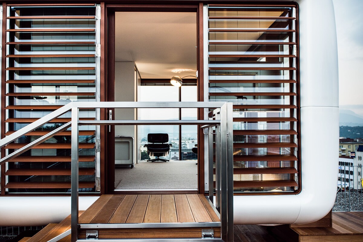A modern interior with a staircase leading to a balcony, a view of the city, and a glimpse of the interior space.