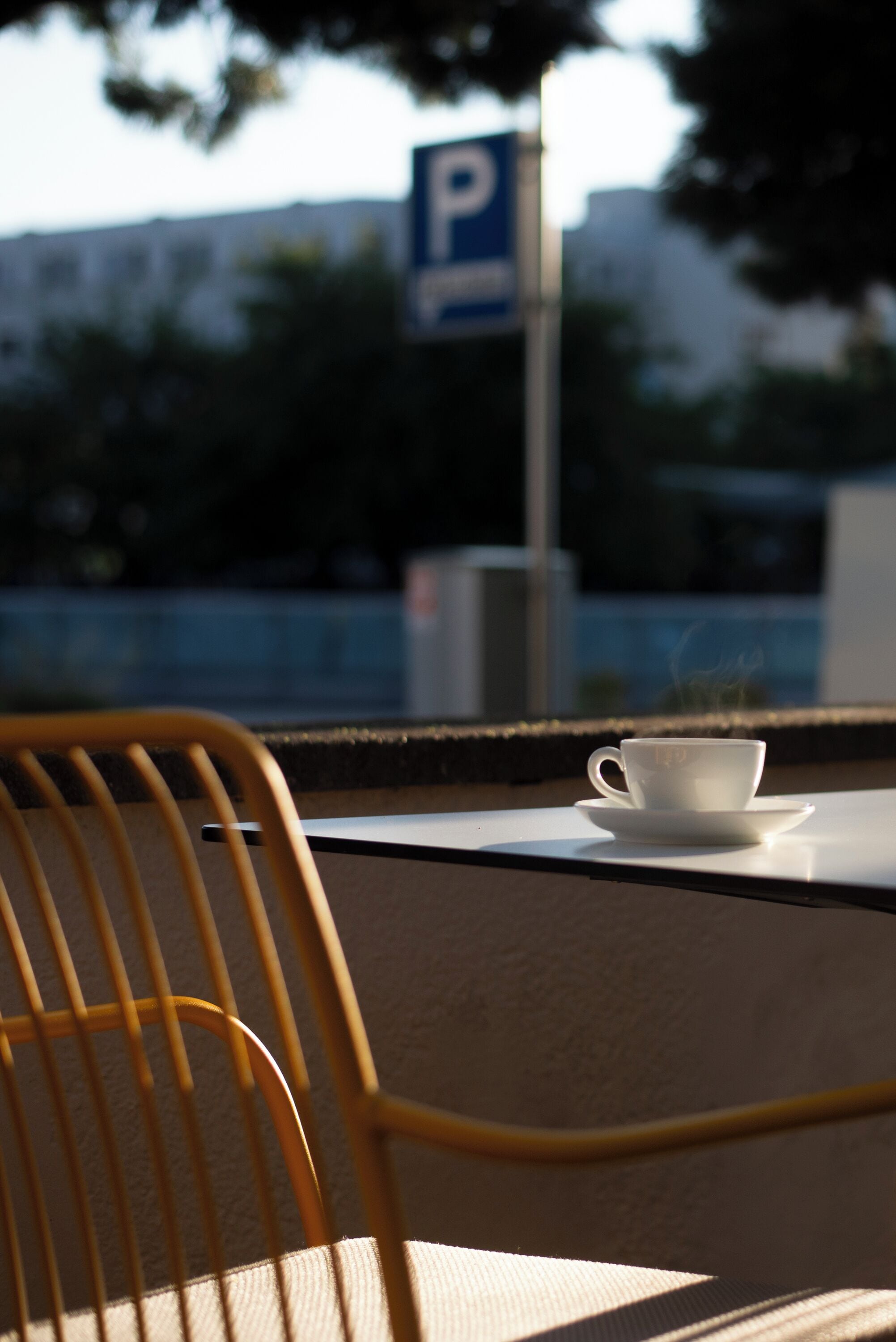 Una taza blanca sobre un platito, sobre una mesa, una silla y un letrero de estacionamiento al fondo.