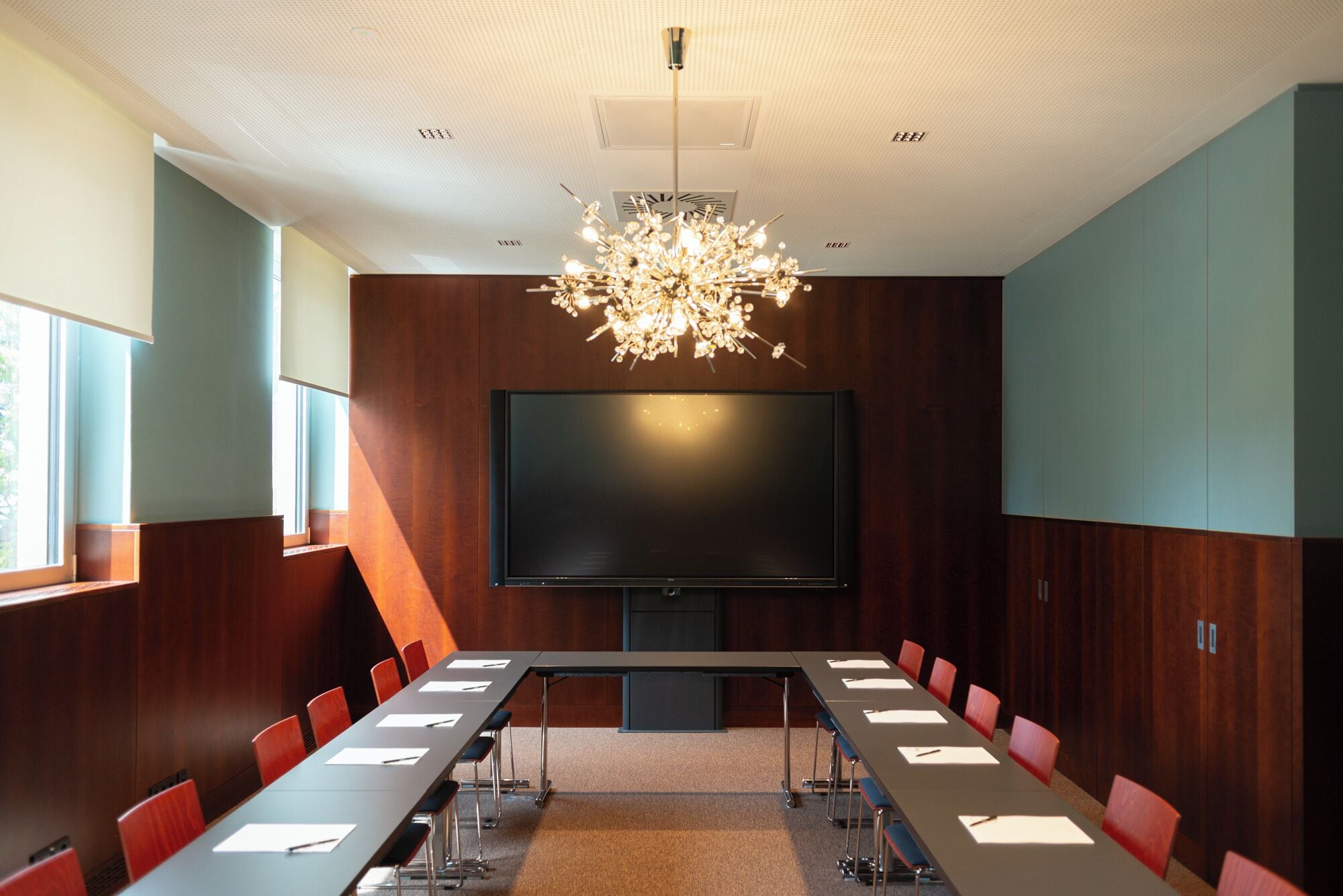 A conference room with a large flat-screen TV, a chandelier, and red chairs.