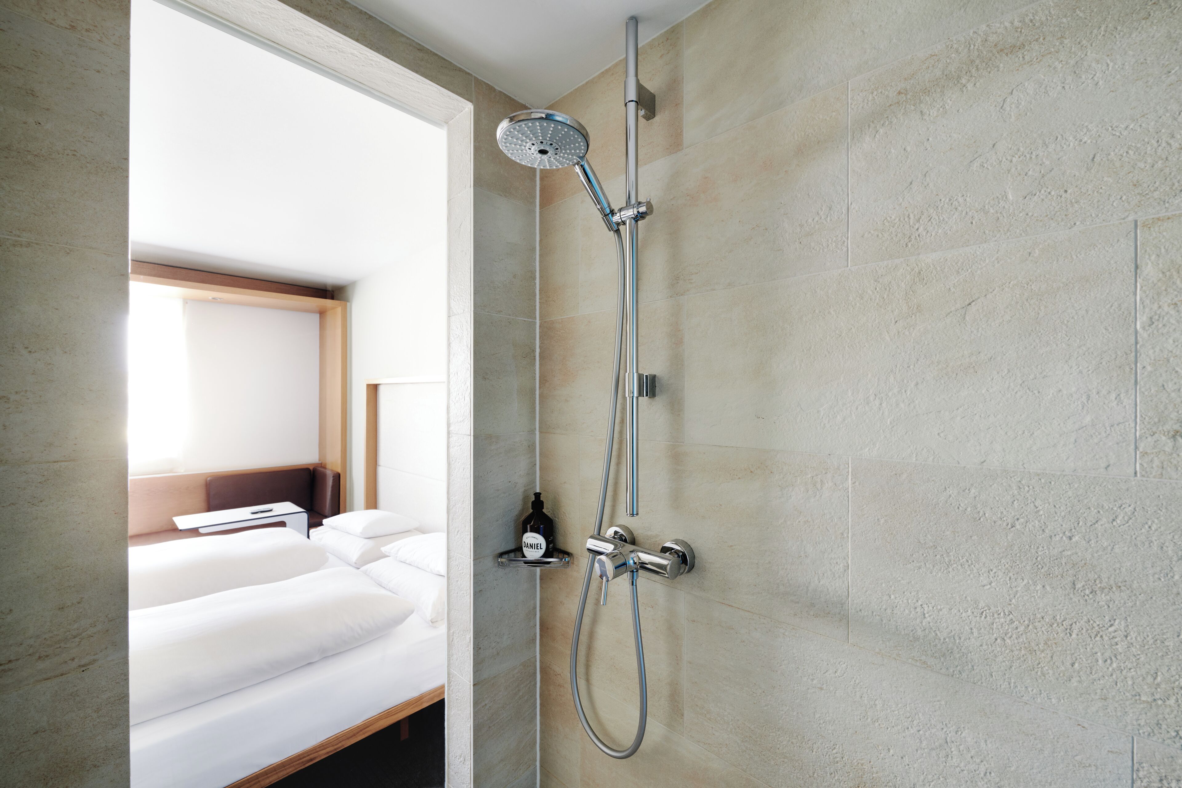 A modern bathroom with a shower, a bed, and a small shelf with toiletries.