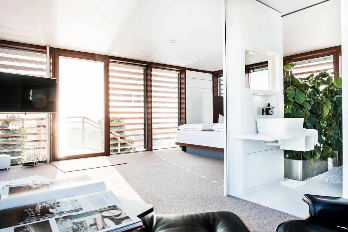 A modern bedroom with a large bed, a bedside table, a white wardrobe, and a large window with blinds.