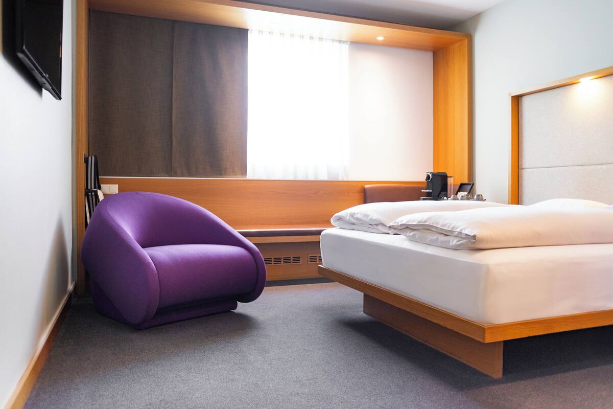 A modern hotel room with a bed, a purple chair, a bedside table, and a window with curtains.
