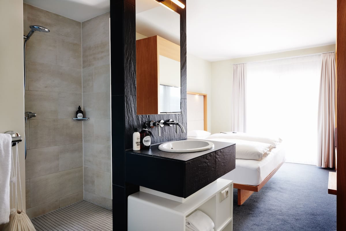 A modern bathroom with a shower, a sink, and a large mirror. There is a bed with white bedding in the adjacent room.