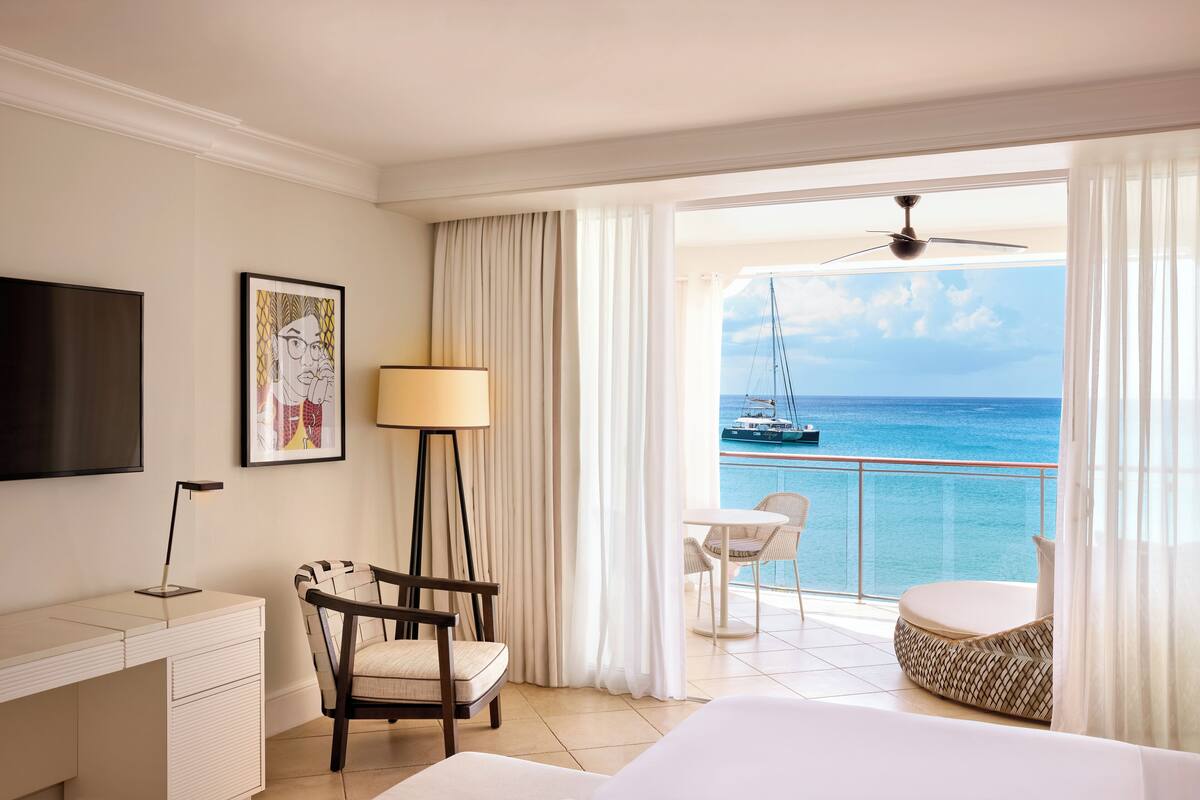 Ein Hotelzimmer mit einem Bett, einem Schreibtisch, einem Sessel, einem Fernseher, einer Lampe und einem Balkon mit Blick auf das Meer und ein Segelboot.