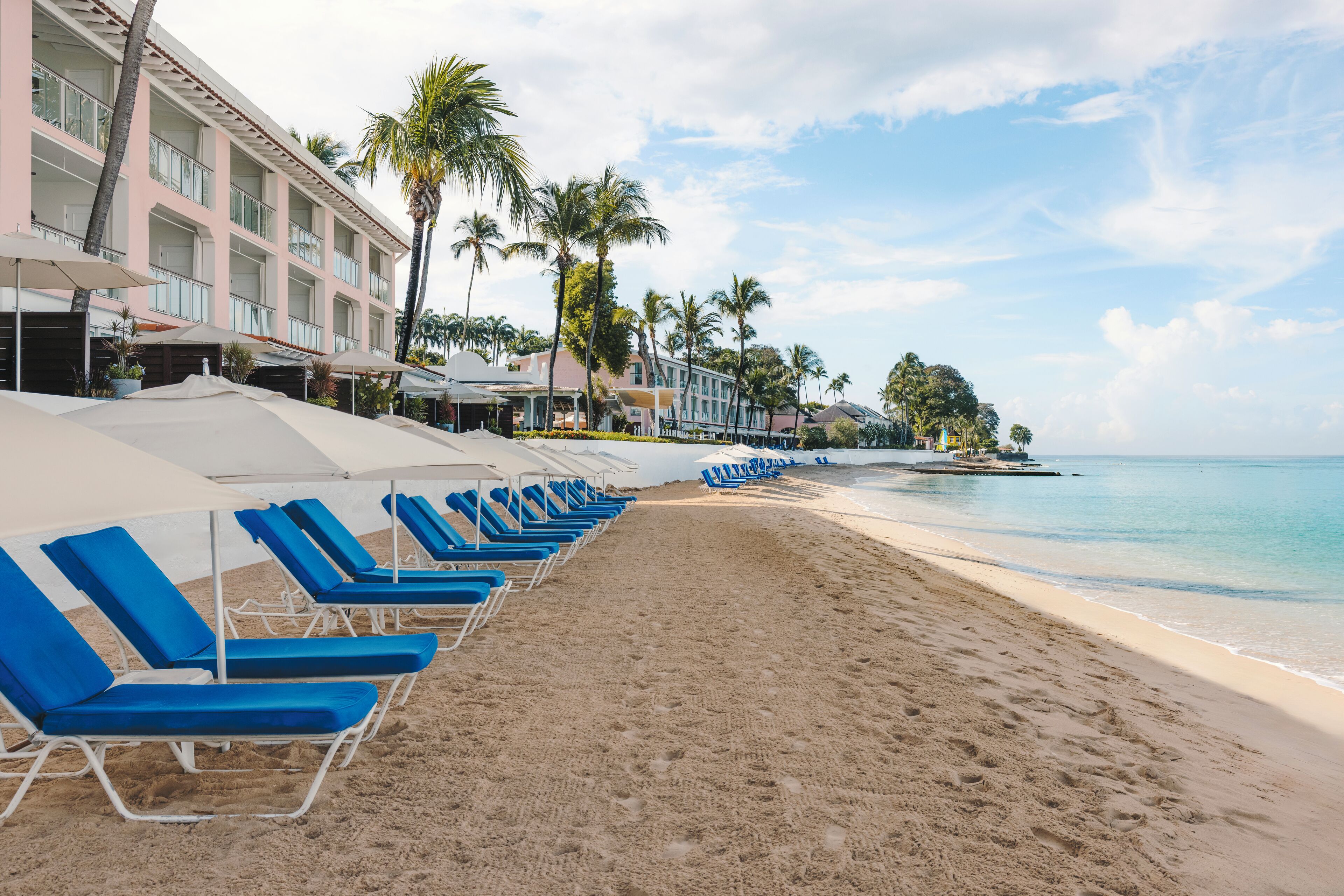 Ein Strand mit blauen Liegestühlen und Sonnenschirmen, ein Hotelgebäude mit Balkonen und Palmen.