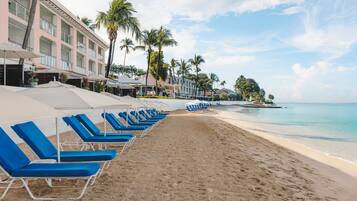 A beach with rows of blue lounge chairs and umbrellas, a hotel building with balconies, and palm trees.